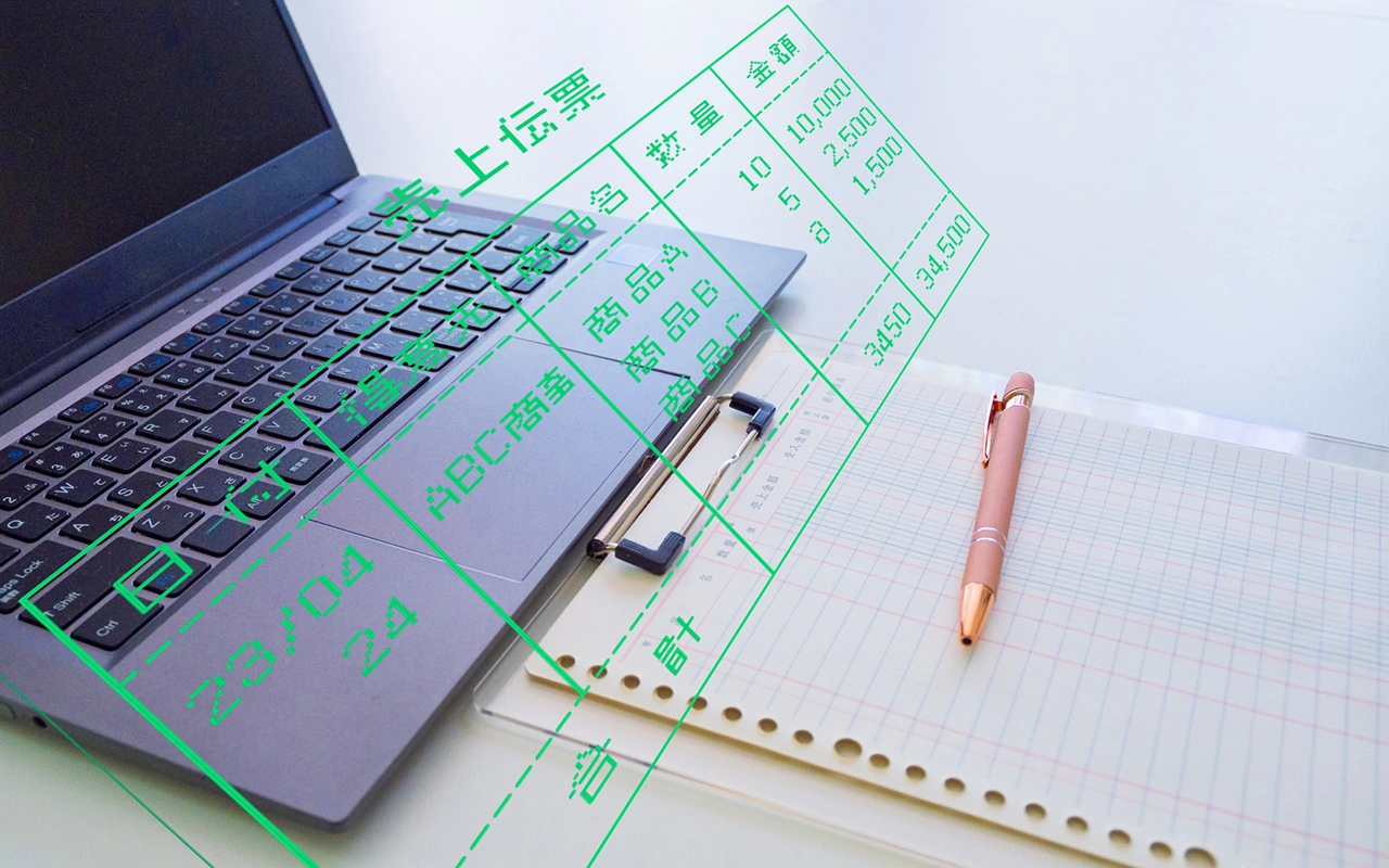 パソコンと会計帳簿に売上伝票がオーバーレイ